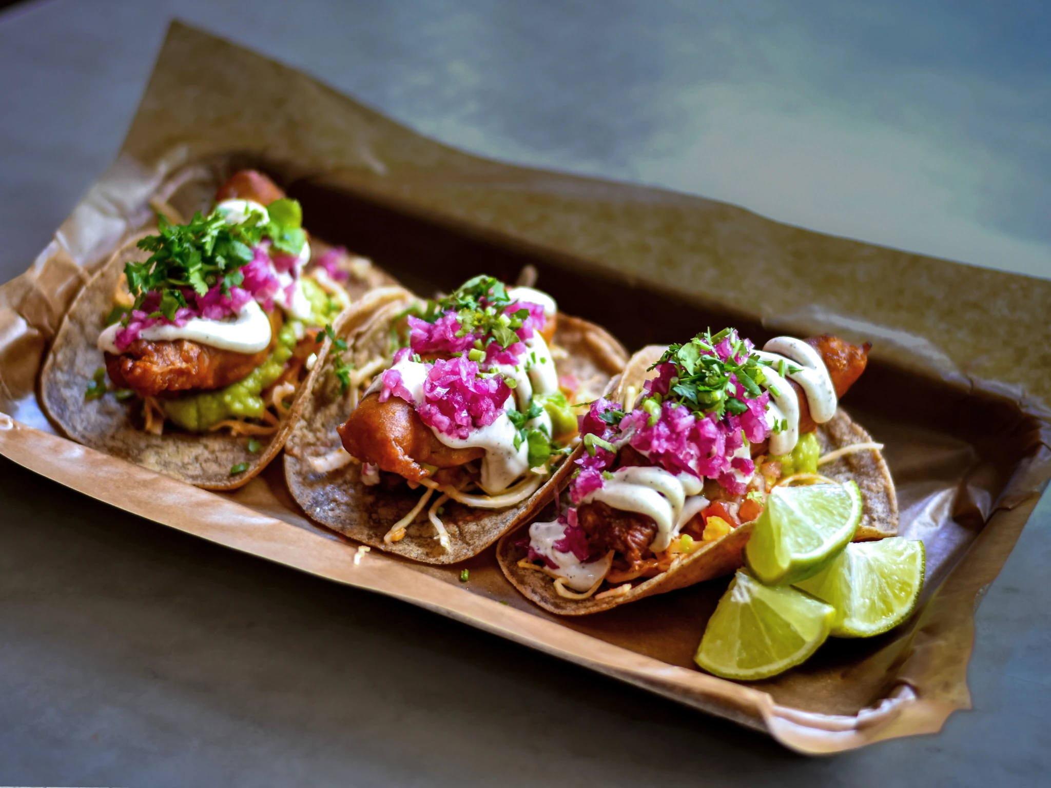 Three loaded tacos filled with fried fish, crema, pickled red onions, and cilantro are served in a paper-lined tray next to lime wedges.