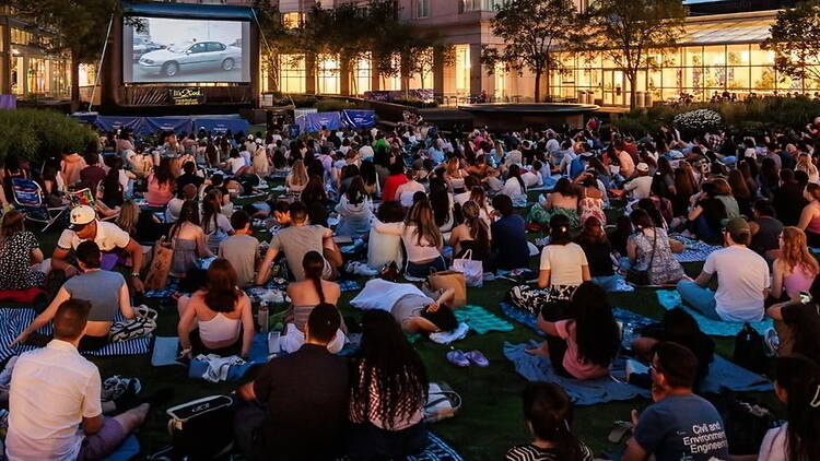 A wide shot of people watching a movie on an outdoor screen in a park. A wide shot of people watching a movie on an outdoor screen in a park.