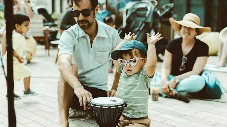 A close-up of a child raising their hands beside a person playing a drum outdoors. A close-up of a child raising their hands beside a person playing a drum outdoors.