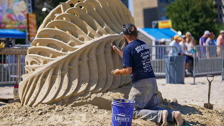 A wide shot of a person carving a large sand sculpture on a beach. A wide shot of a person carving a large sand sculpture on a beach.