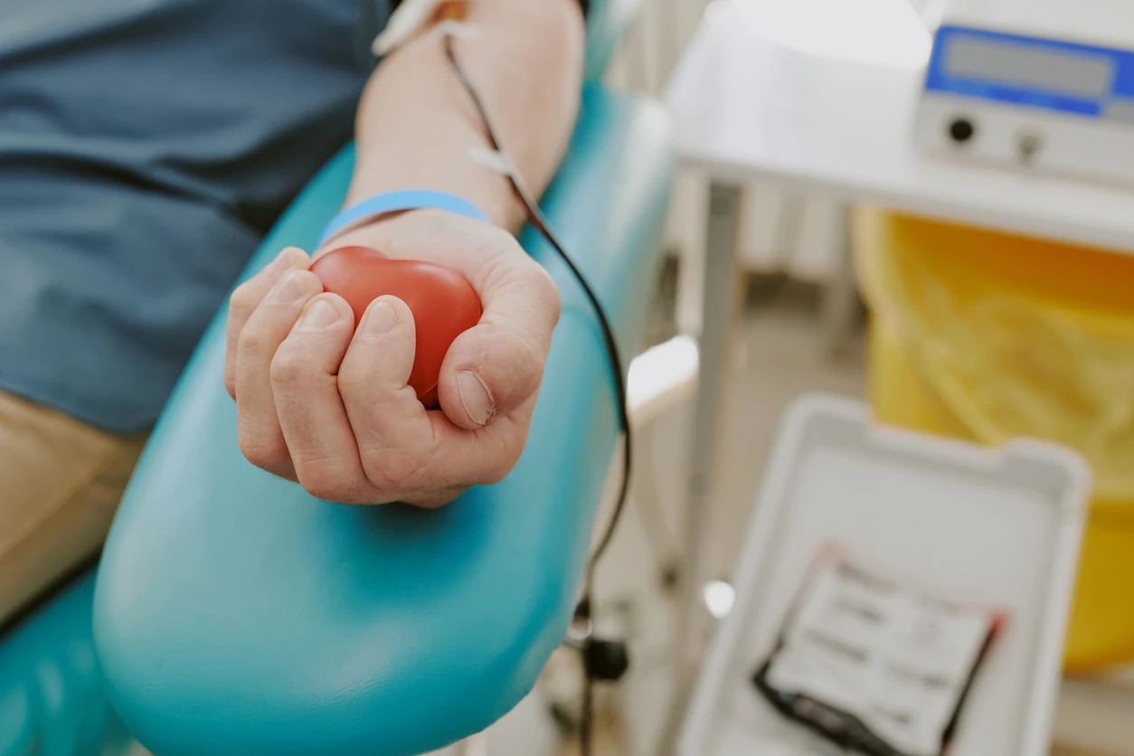 Caucasian middle aged man donating blood, arm resting on medical chair, hand squeezing red stress ball, blood collection tube attached, blood donation bag visible in clinical setting
