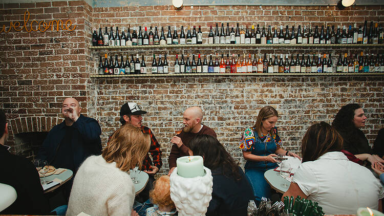 men and women drink wine and converse with brick wall and wine bottles in the background
