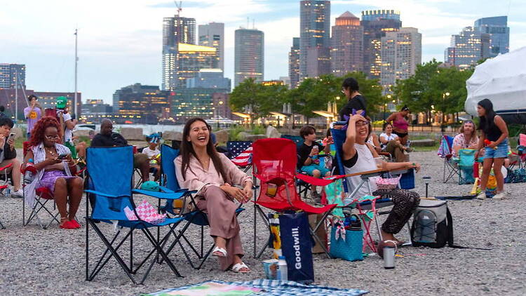 A wide shot of people relaxing on chairs at an outdoor waterfront event with a city skyline backdrop A wide shot of people relaxing on chairs at an outdoor waterfront event with a city skyline backdrop