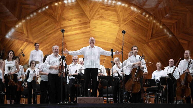 A wide shot of an orchestra performing with a conductor on stage in a wooden concert hall. A wide shot of an orchestra performing with a conductor on stage in a wooden concert hall.