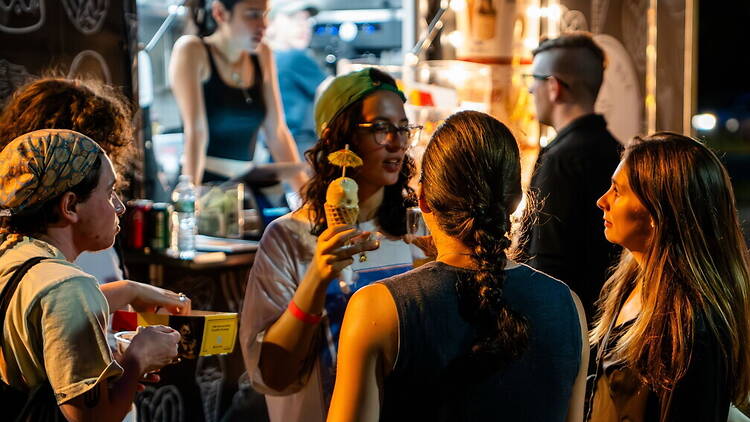 A wide shot of people chatting around a food truck at an outdoor evening event. A wide shot of people chatting around a food truck at an outdoor evening event.