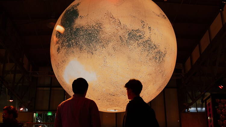 A wide shot of two people viewing a large moon exhibit in a museum.