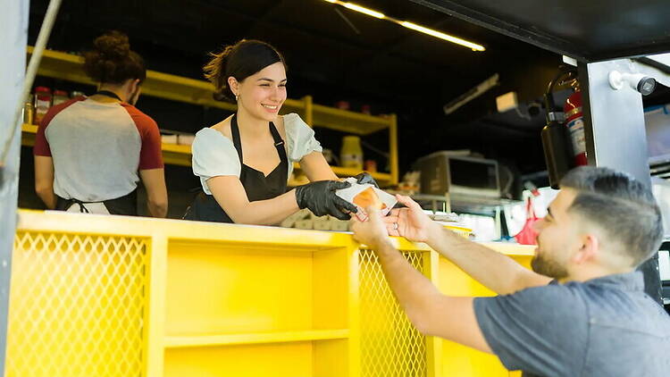 A wide shot of a food truck worker handing a wrapped meal to a customer at a food truck. A wide shot of a food truck worker handing a wrapped meal to a customer at a food truck.
