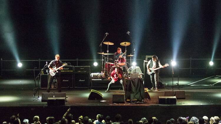 A wide shot of a rock band performing on a brightly lit stage before an audience. A wide shot of a rock band performing on a brightly lit stage before an audience.