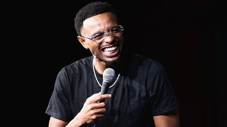 Close-up of a comedian laughing while holding a microphone on a dark stage. Close-up of a comedian laughing while holding a microphone on a dark stage.