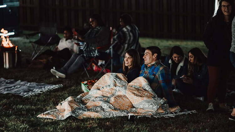 A wide shot of people sitting on blankets at an outdoor gathering. A wide shot of people sitting on blankets at an outdoor gathering.