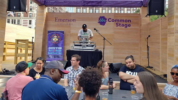 A wide shot of a DJ performing on an outdoor stage with people seated in front. A wide shot of a DJ performing on an outdoor stage with people seated in front.