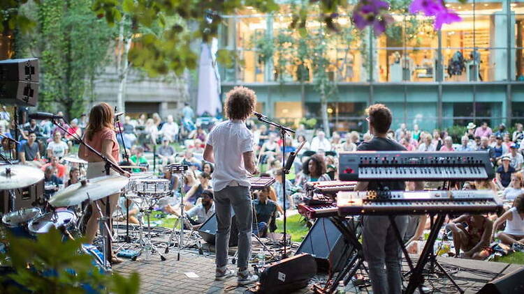 A wide shot of a band performing on an outdoor stage before a seated audience A wide shot of a band performing on an outdoor stage before a seated audience
