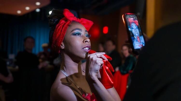 Close-up of a performer applying lipstick while facing a smartphone camera at an indoor event. Close-up of a performer applying lipstick while facing a smartphone camera at an indoor event.