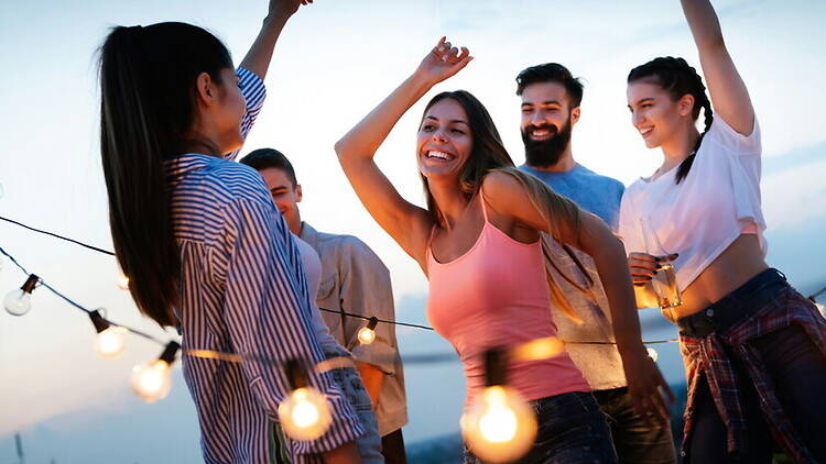A wide shot of people dancing together on a rooftop under string lights at sunset. A wide shot of people dancing together on a rooftop under string lights at sunset.