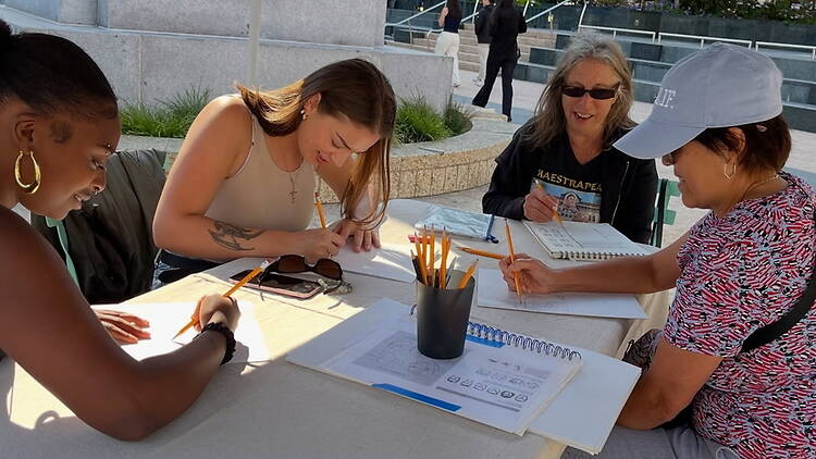 A wide shot of four people drawing together at an outdoor table. A wide shot of four people drawing together at an outdoor table.