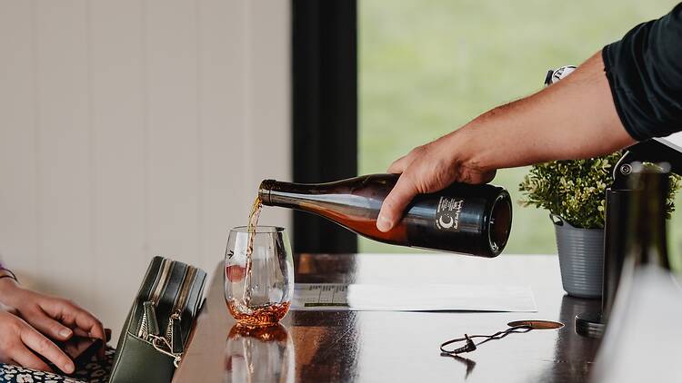 Comedy in the Vines person pours wine into a glass for another person
