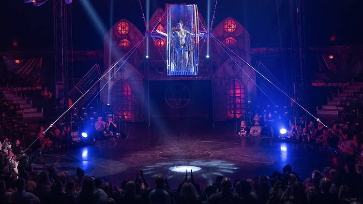 A wide shot of an aerial performer suspended above a circus stage in front of an audience.