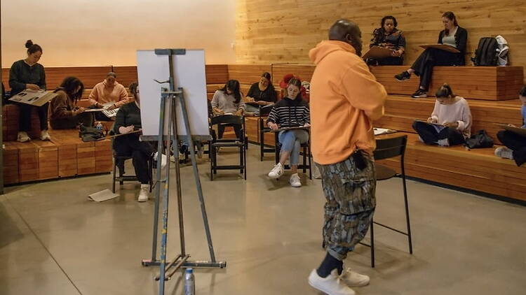 A wide shot of people sketching while an instructor speaks in a tiered classroom. A wide shot of people sketching while an instructor speaks in a tiered classroom.