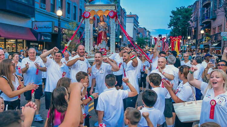 A wide shot of people celebrating during a street procession carrying a religious statue through a city. A wide shot of people celebrating during a street procession carrying a religious statue through a city.