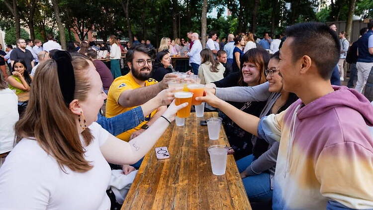 A wide shot of people raising drinks together at picnic tables in an outdoor gathering. A wide shot of people raising drinks together at picnic tables in an outdoor gathering.