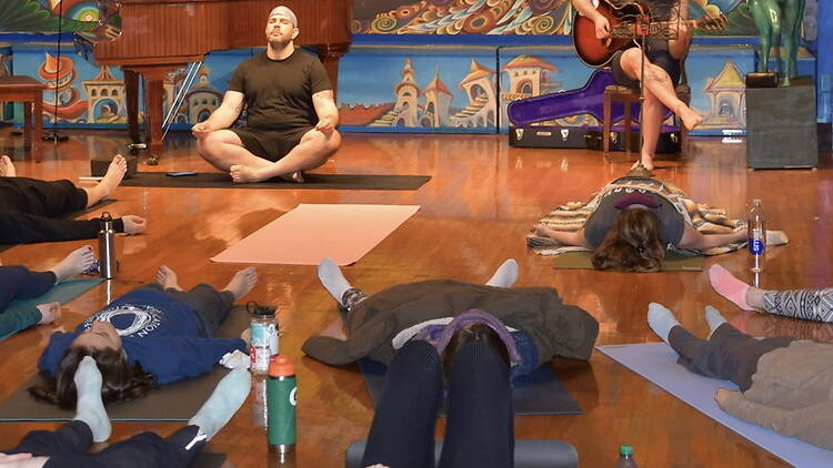 A wide shot of people lying on yoga mats while an instructor leads a session indoors. A wide shot of people lying on yoga mats while an instructor leads a session indoors.