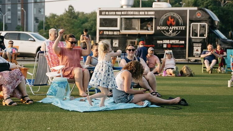 A wide shot of people enjoying a picnic on a lawn with a food truck in the background. A wide shot of people enjoying a picnic on a lawn with a food truck in the background.