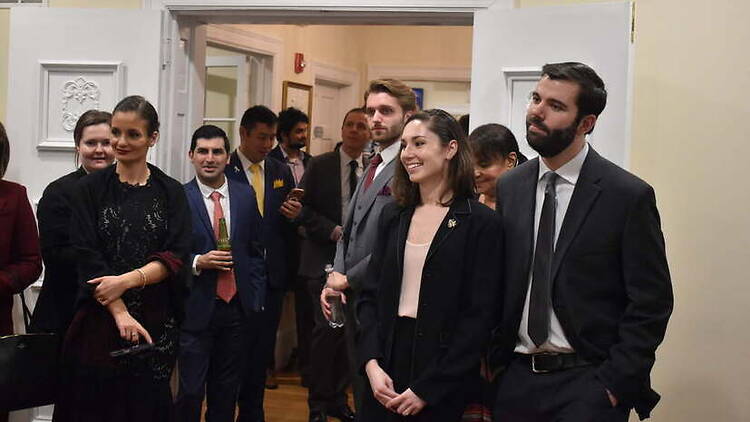 A wide shot of formally dressed guests gathering in a reception room. A wide shot of formally dressed guests gathering in a reception room.