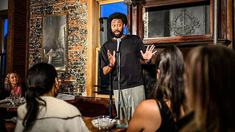 A wide shot of a comedian performing on stage in front of an audience at an indoor venue. A wide shot of a comedian performing on stage in front of an audience at an indoor venue.