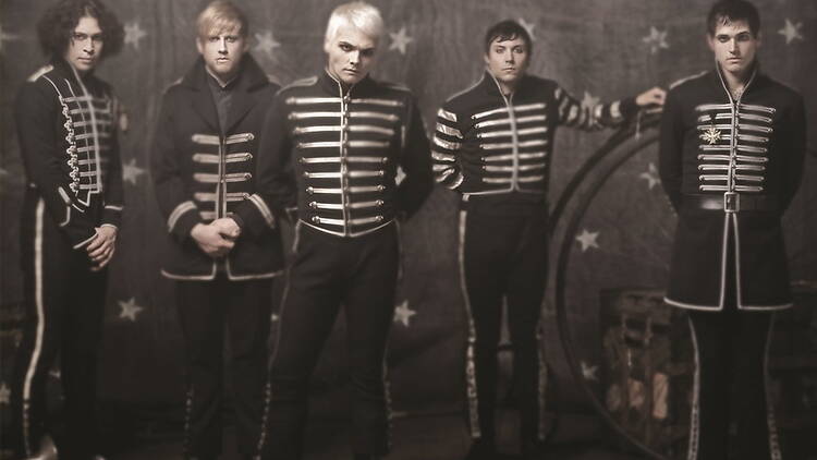 A wide shot of five band members posing in matching military-style jackets before a vintage circus backdrop A wide shot of five band members posing in matching military-style jackets before a vintage circus backdrop