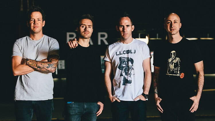 A wide shot of four band members posing together in an indoor venue A wide shot of four band members posing together in an indoor venue