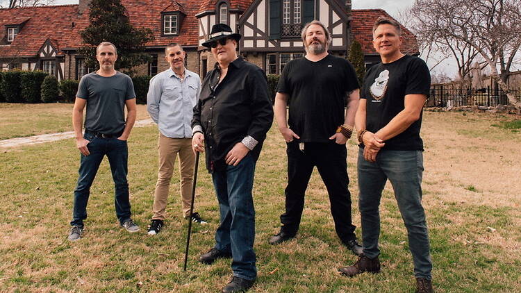 A wide shot of five band members posing on a lawn in front of a Tudor-style building A wide shot of five band members posing on a lawn in front of a Tudor-style building