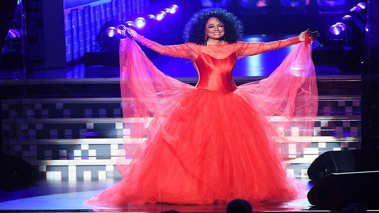 A wide shot of a performer singing on stage in a flowing red gown under concert lights A wide shot of a performer singing on stage in a flowing red gown under concert lights
