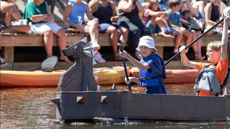 A wide shot of two children paddling a cardboard boat on a lake during an outdoor event. A wide shot of two children paddling a cardboard boat on a lake during an outdoor event.