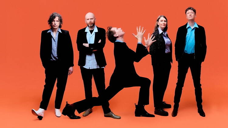 A wide shot of five people posing against an orange studio background. A wide shot of five people posing against an orange studio background.