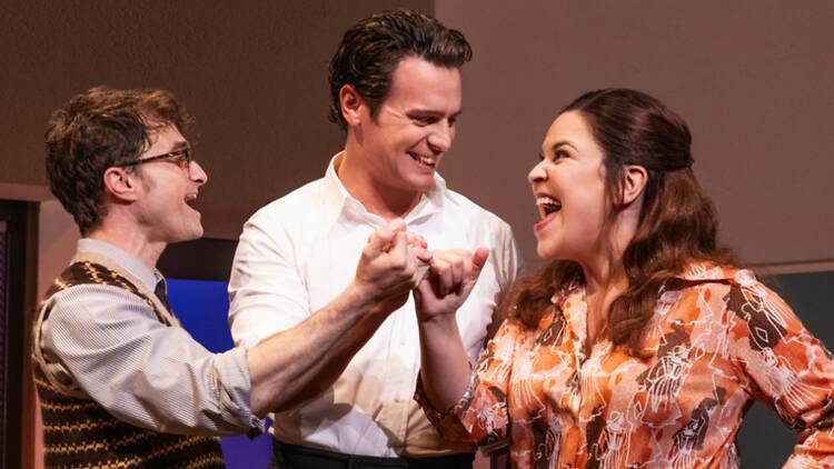 A close-up of three actors smiling and holding hands on a theater stage. A close-up of three actors smiling and holding hands on a theater stage.