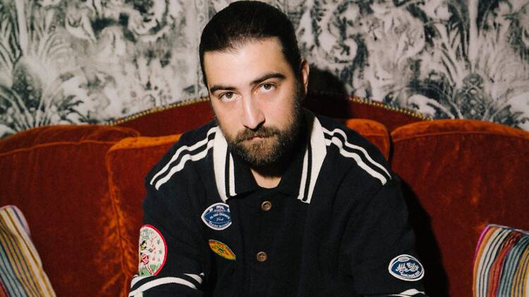 Close-up of a bearded man sitting on an orange sofa in front of patterned wallpaper. Close-up of a bearded man sitting on an orange sofa in front of patterned wallpaper.