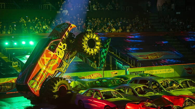A wide shot of a monster truck performing a wheelie over crushed cars in an indoor arena. A wide shot of a monster truck performing a wheelie over crushed cars in an indoor arena.
