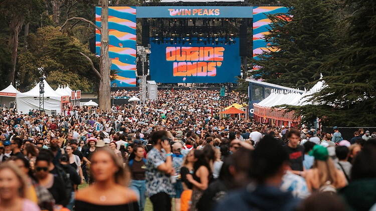 A wide shot of a crowd gathering in front of a festival stage at an outdoor park. A wide shot of a crowd gathering in front of a festival stage at an outdoor park.
