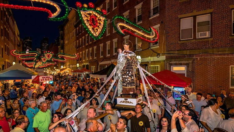A wide shot of a crowd carrying a religious statue through a decorated street at night. A wide shot of a crowd carrying a religious statue through a decorated street at night.