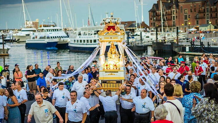 A wide shot of people carrying a religious statue along a waterfront promenade. A wide shot of people carrying a religious statue along a waterfront promenade.