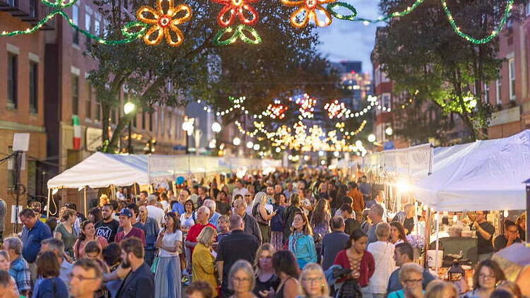 A wide shot of people browsing vendor booths at a street festival beneath decorative lights in a city street. A wide shot of people browsing vendor booths at a street festival beneath decorative lights in a city street.