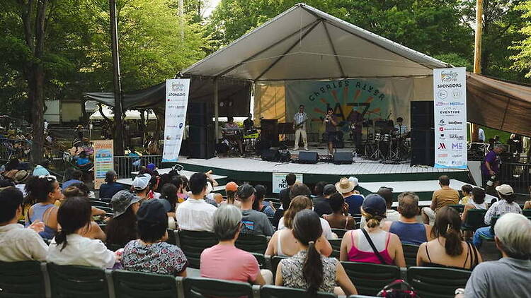 A wide shot of a band performing on an outdoor stage before a seated audience in a park. A wide shot of a band performing on an outdoor stage before a seated audience in a park.