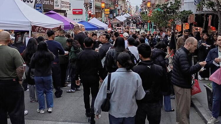 A wide shot of people walking through a crowded street market lined with vendor tents. A wide shot of people walking through a crowded street market lined with vendor tents.