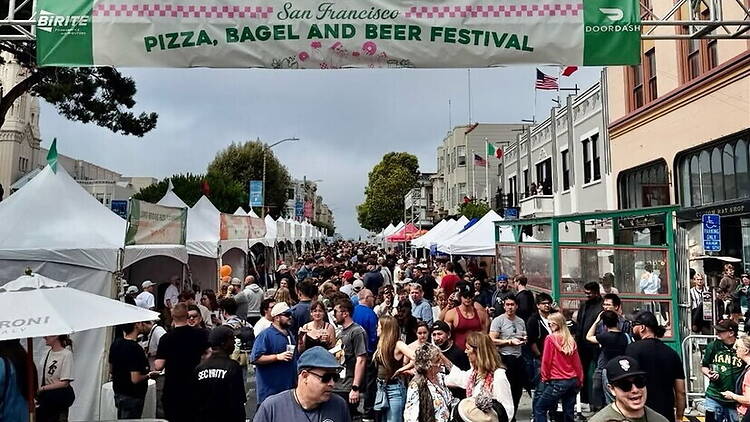 A wide shot of people gathering at a street festival beneath a festival banner.