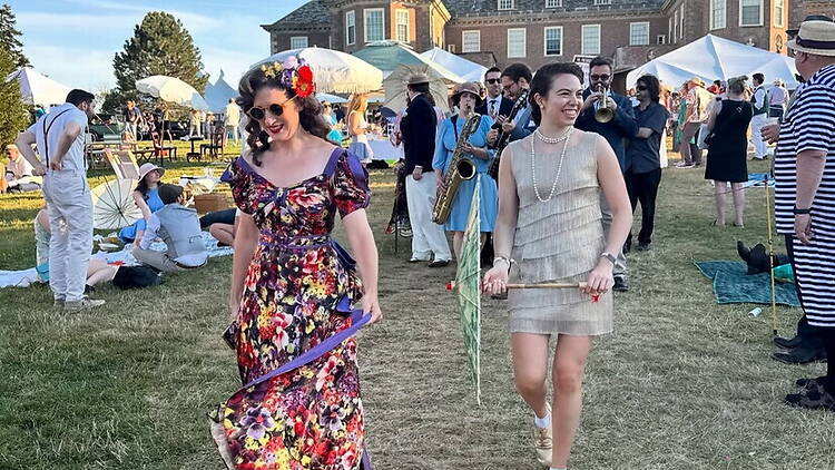 A wide shot of people walking across a festival lawn with a live band performing nearby. A wide shot of people walking across a festival lawn with a live band performing nearby.