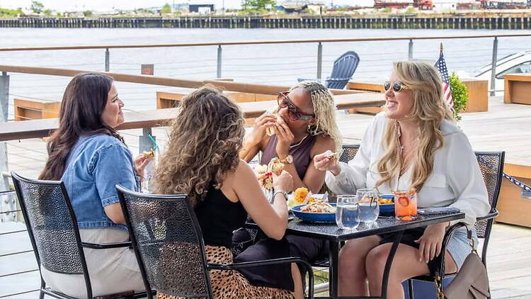 A wide shot of people eating and chatting at an outdoor restaurant by the waterfront. A wide shot of people eating and chatting at an outdoor restaurant by the waterfront.