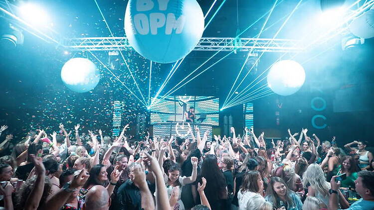 A wide shot of a crowd dancing under stage lights at an indoor venue. A wide shot of a crowd dancing under stage lights at an indoor venue.