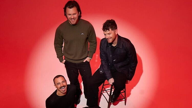 A wide shot of three people smiling and posing in a photo studio with a red backdrop. A wide shot of three people smiling and posing in a photo studio with a red backdrop.
