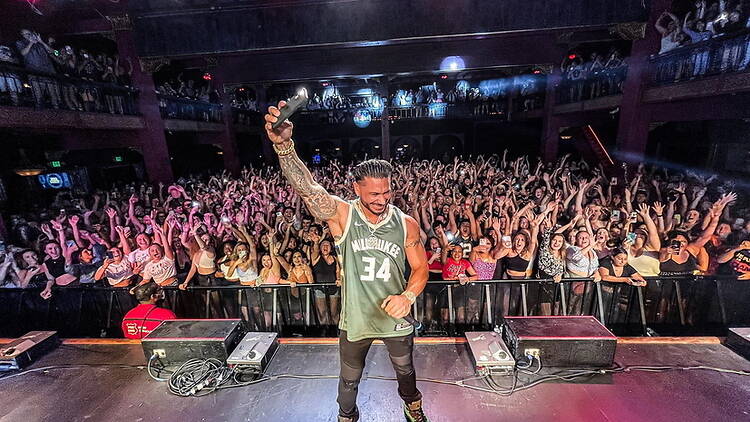 A wide shot of a performer raising a microphone to a cheering crowd on a concert stage. A wide shot of a performer raising a microphone to a cheering crowd on a concert stage.