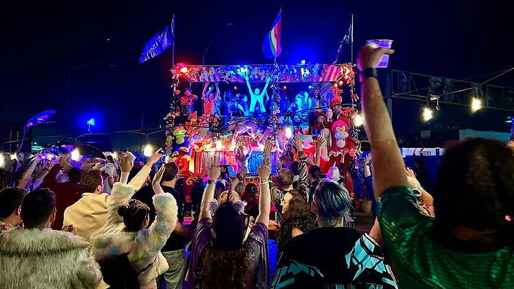 A wide shot of a crowd cheering in front of a decorated parade float at a nighttime festival. A wide shot of a crowd cheering in front of a decorated parade float at a nighttime festival.
